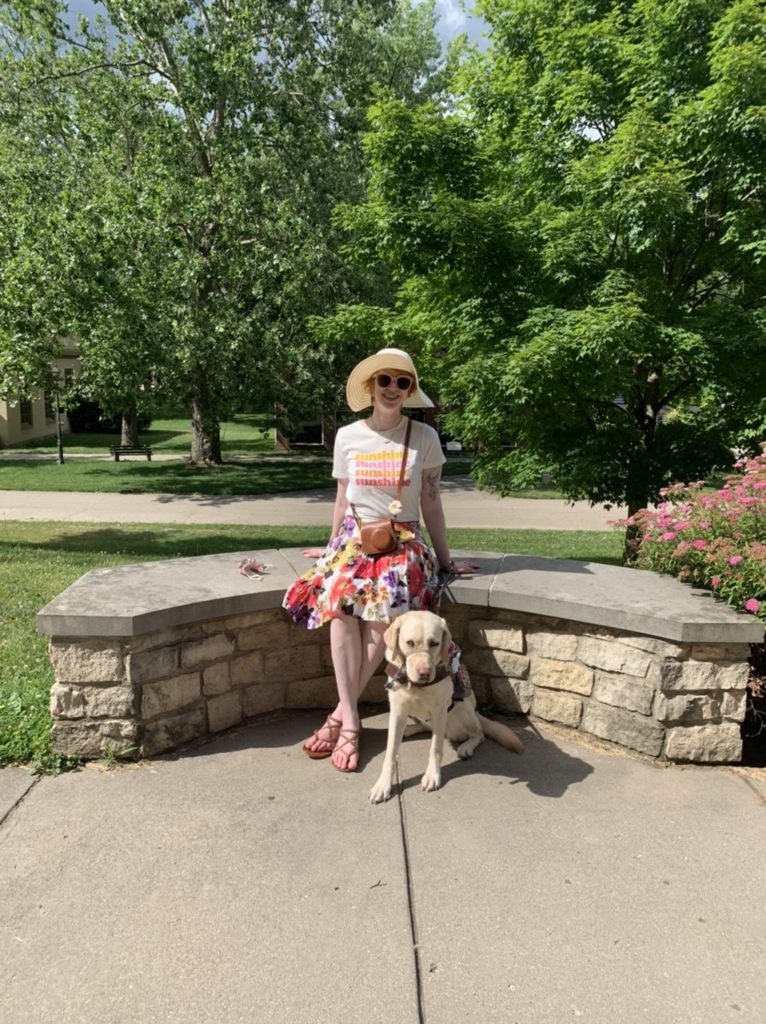 cassandra outside with guide dog romana sitting on a stone bench. cassandra wears a bright floral skirt, brown strappy sandals, a white graphic tee with the word "sunshine" four times, and a white wide brimmed hat