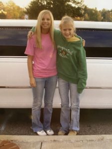 casey and sister lynley outside in front of a white car. casey wears a green aeropostale hoodie with a monkey on it and light wash jeans. caseys blond hair is frizzy. her sister wears a bright pink tee and light wash jeans