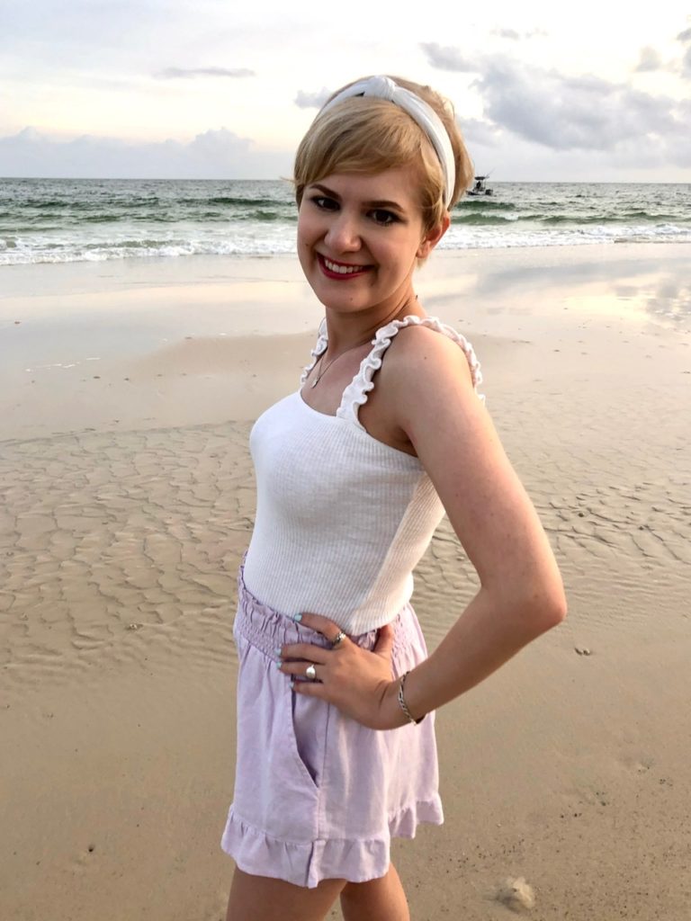 casey smiling at the beach, sporting a white halter top and lavender high waisted shorts. she has a pixie cut with a white headband