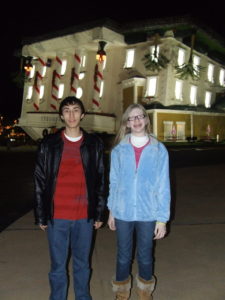 cassandra and brother carlos standing outside at night. cassandra wears a white turtleneck under a bright pink tee, with a fuzzy light blue north face jacket on top. her hair is blond and frizzy. her brother wears a red american eagle tee