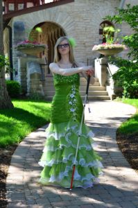 cassandra with long blonde hair posing with her hands on top of her cane with it outstretched. she wears an elaborate green and white mermaid style prom dress. the dress has sparkly rhinestones, green and white flowers and feathers, and big ruffles at the bottom