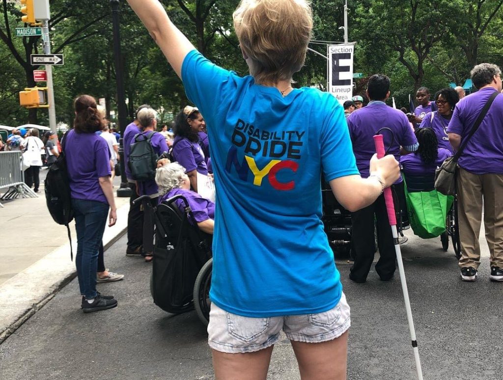 casey, with her back to the camera, wears a blue tshirt that says "disability pride NYC.” her short blond hair is visible, and she has her left hand thrown up while her right hand holds her pink cane in a power stance