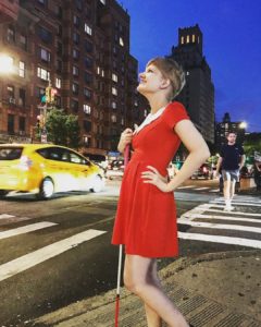 short haired blonde casey poses on a street corner in manhattan. she wears a red dress with a large whit collar and stands facing left, holding her cane in one hand and with the other hand on her hip. behind her you can see the city with a yellow taxi and a pedestrian