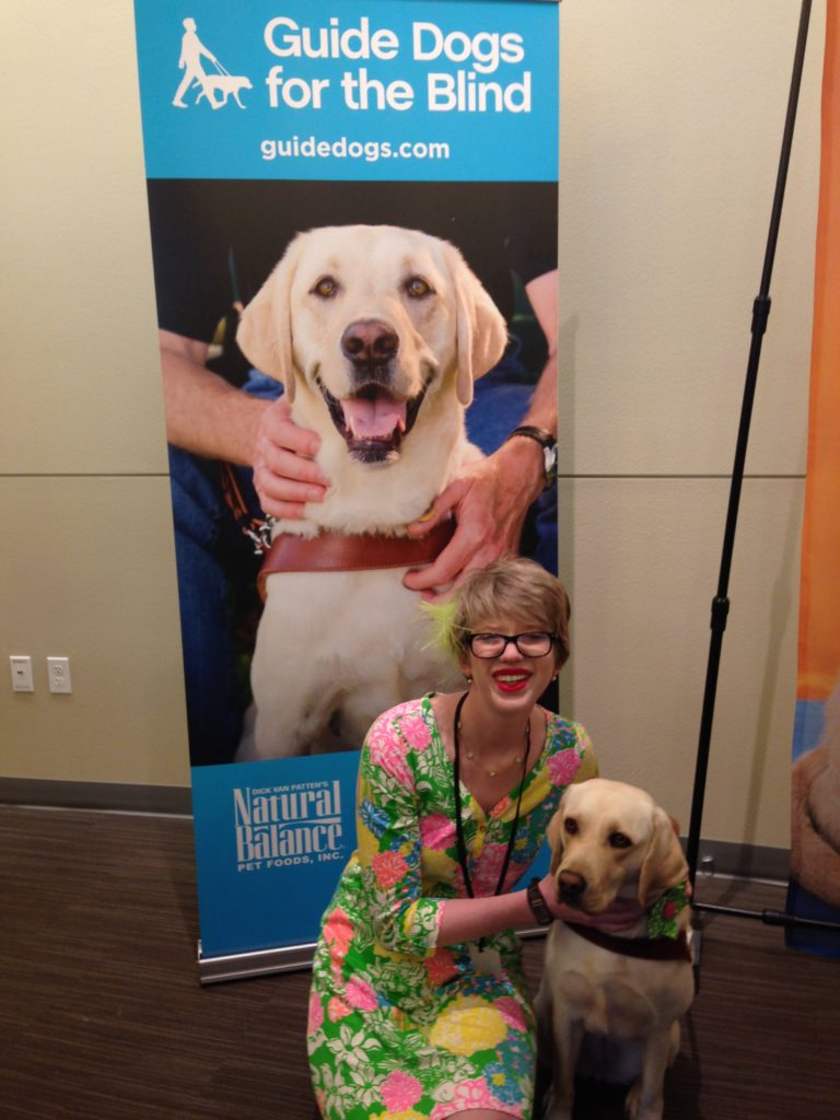 short haired, blonde cassandra kneels next to yellow lab romana in harness. cassandra wears a colorful floral dress and romana has a green flower in her collar. cass looks ecstatic and ro is focused