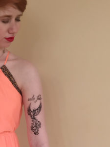 Cassandra posing against a cream colored wall and only her top left is visible. her bicep tattoo is the focus, which is a phoenix drawn of lines and swirls as it rises. "Sola fide" is written in script above