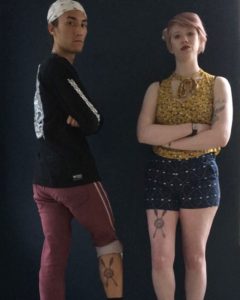 Cassandra right and older brother carlos left posing with their matching tattoos against a dark wall. both look serious with crossed arms. carlos looks over his shoulder to face the camera as his rolled pant leg revels the back of his calf where his tattoo is. cassandra wears shorts and her thigh tattoo is visible 