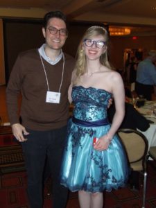 cassandra standing with a white adult male, Dr. Cohen. cassandra wears a blue homecoming-style dress and has long blonde hair, and Dr. Cohen wears a brown sweater and jeans with dark hair and toirtoseshell glasses