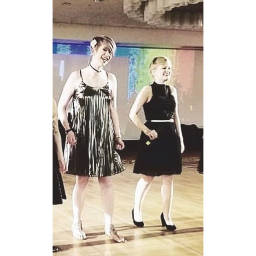 cassandra and casey performing at the HPS conference. Cassandra wears a silver dress and Casey wears a black one. they both hold microphones