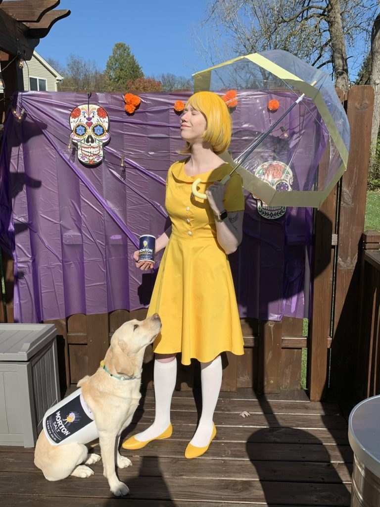 Cassandra is dressed as the Morton Salt girl. She's wearing a yellow dress and yellow wig. She is holding an umbrella in one hand and a salt container in the other. Romana sits in front of her with the salt container logo wrapped around her