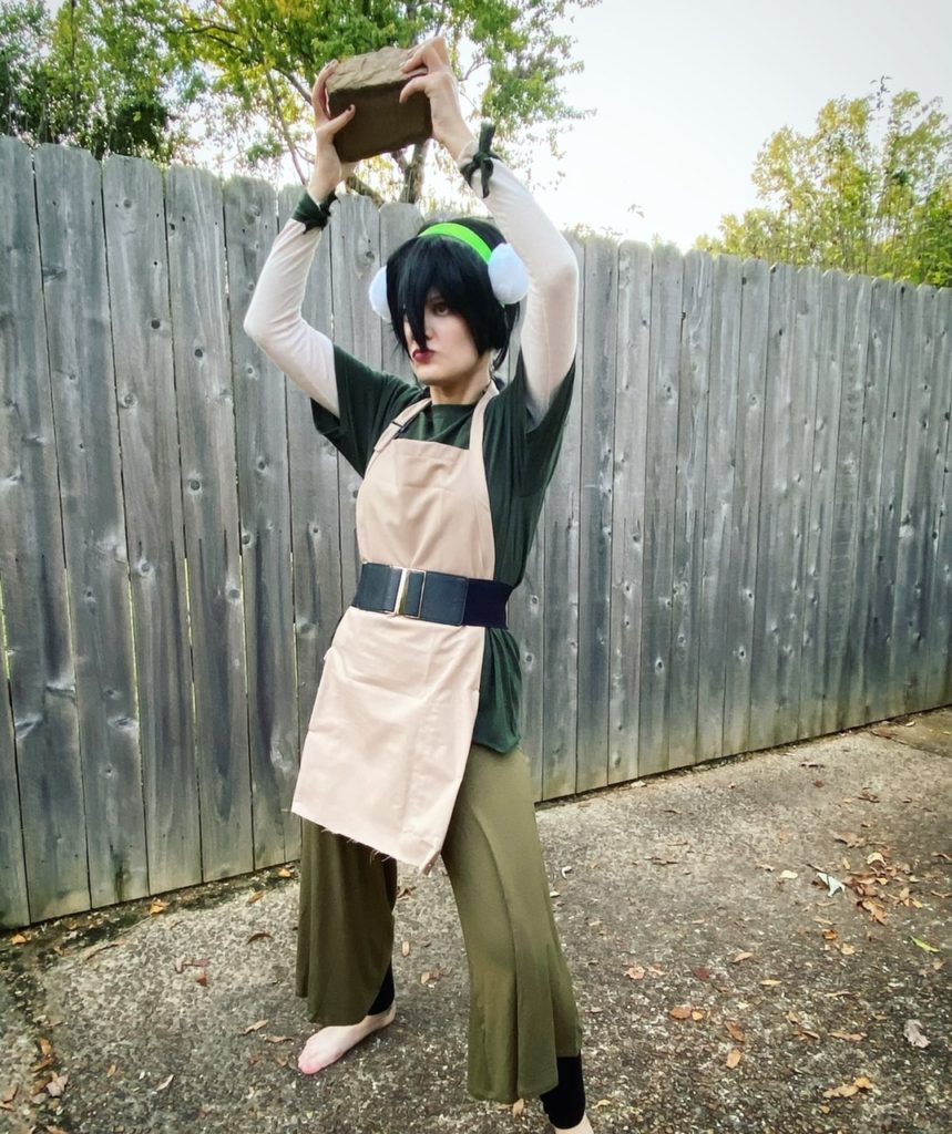 Casey is dressed as Toph from Avatar the last Airbender. She is wearing a green outfit, a black wig, and is holding a rock over her head