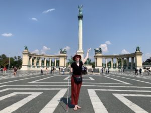 cass standing in a plaza with statues of famous communist leaders in Budapest. she wears red pants, a black top, and a red felt hat. she carries her cane and raises one hand aloft