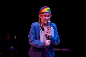 casey onstage dressed as a classic "geek" wearing large, red glasses, a multicolored knit hat, and braids. she is in character and grabs a prop mic with both hands and curls her lips