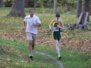 cassandra in a green and yellow uniform and black baseball cap running cross country alongside a sighted guide in the fall