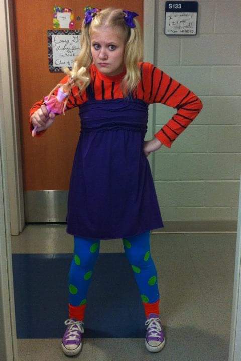 teenage casey dressed as angelica pickles. she wears her hair in pigtails, an orange longlseeve, purple jumper dress, blue leggings and converse with orange socks. she pouts and puts her hand on her hip and holds a cynthia doll with the other hand