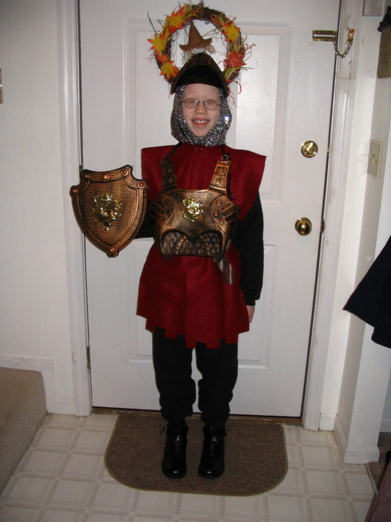 young cass (8) dressed as a knight with a burgundy dress and plastic armor and shield with fake chainmail around her neck and head