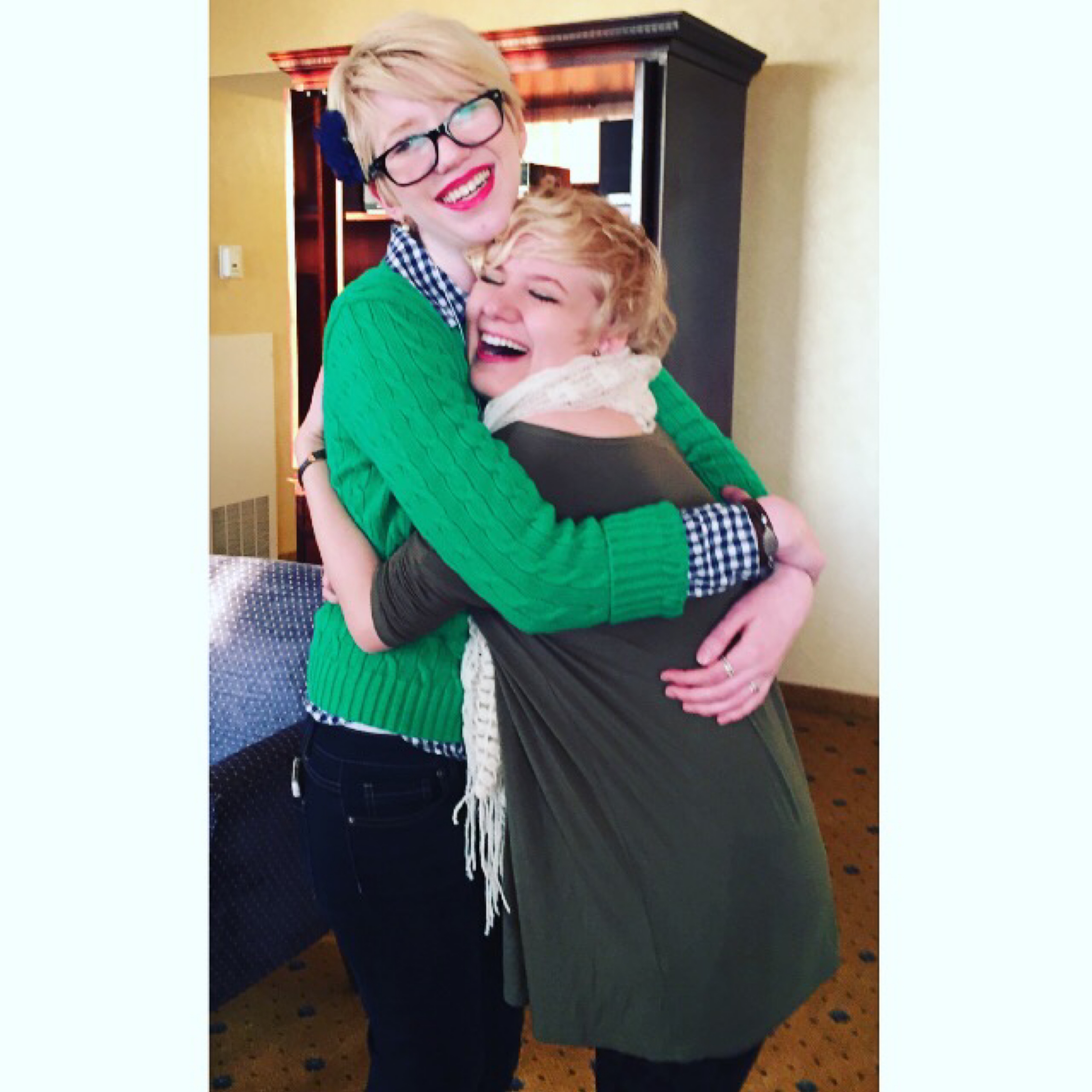 Photo of case and cass hugging in a hotel room. cass has a green cable knit sweater and glasses, and smiles widely as she embraces case, who’s shorter and laughs and smiles while pressed agains cass’ chest. case wears a brown dress and white scarf