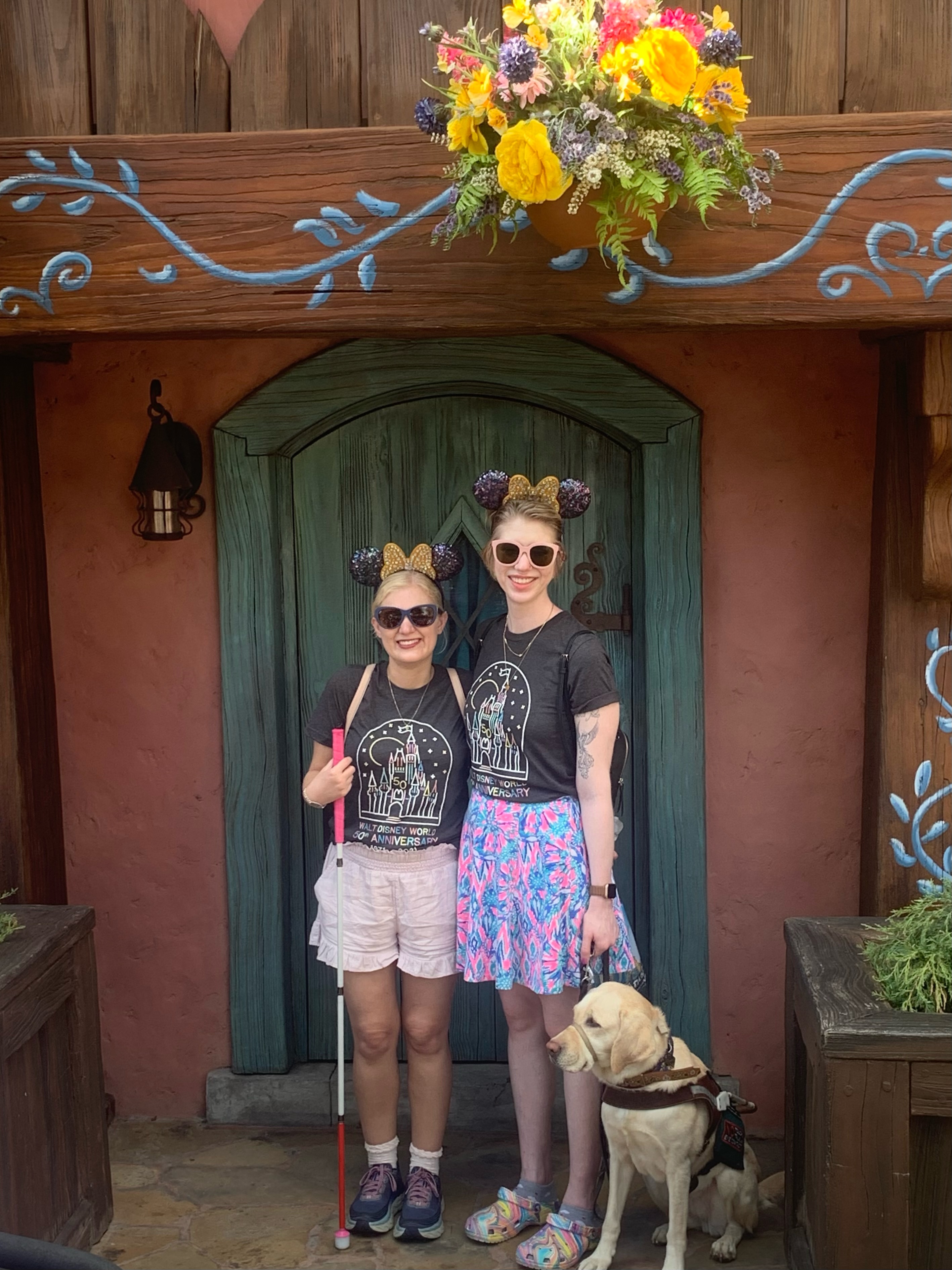 photo of casey and cassandra in disney world in front of a wood painted wall. they wear matching gray tshirts and sunglasses, and both s,i le at the camera. cass’ yellow lab guide dog romana looks off to the left