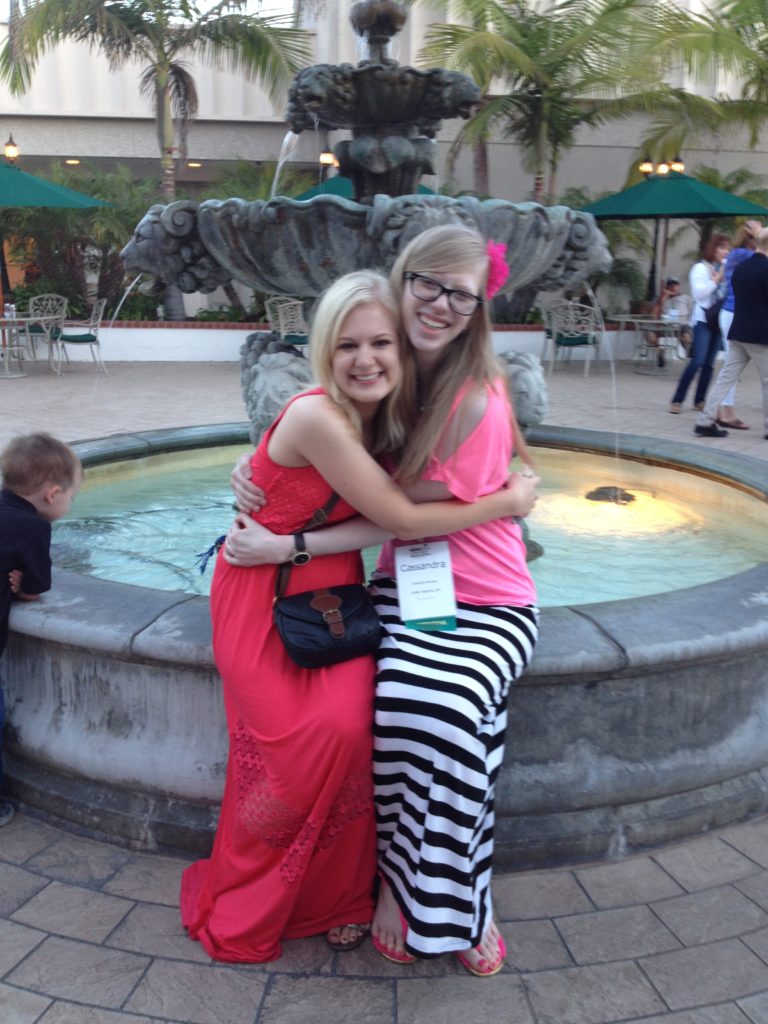 young casey and cassandra sitting on the edge of a concrete fountain with lights. they embrace and smile at they camera. casey wears a red maxi dress and shoulder-length light blonde hair, and cassandra wears a pink top with a black and white striped maxi skirt with her long, medium blonde hair