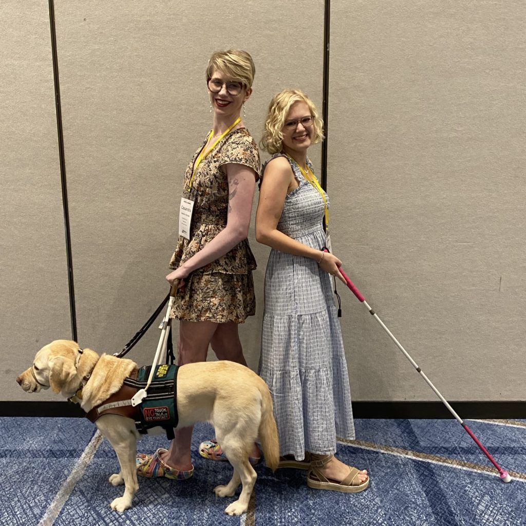 case and cass stand back to back, posing like their cover art. cassandra wears a beige floral mini dress and holds romana’s harness handle. casey wears a long, pale blue dress and extends her pink handled came out in front of her