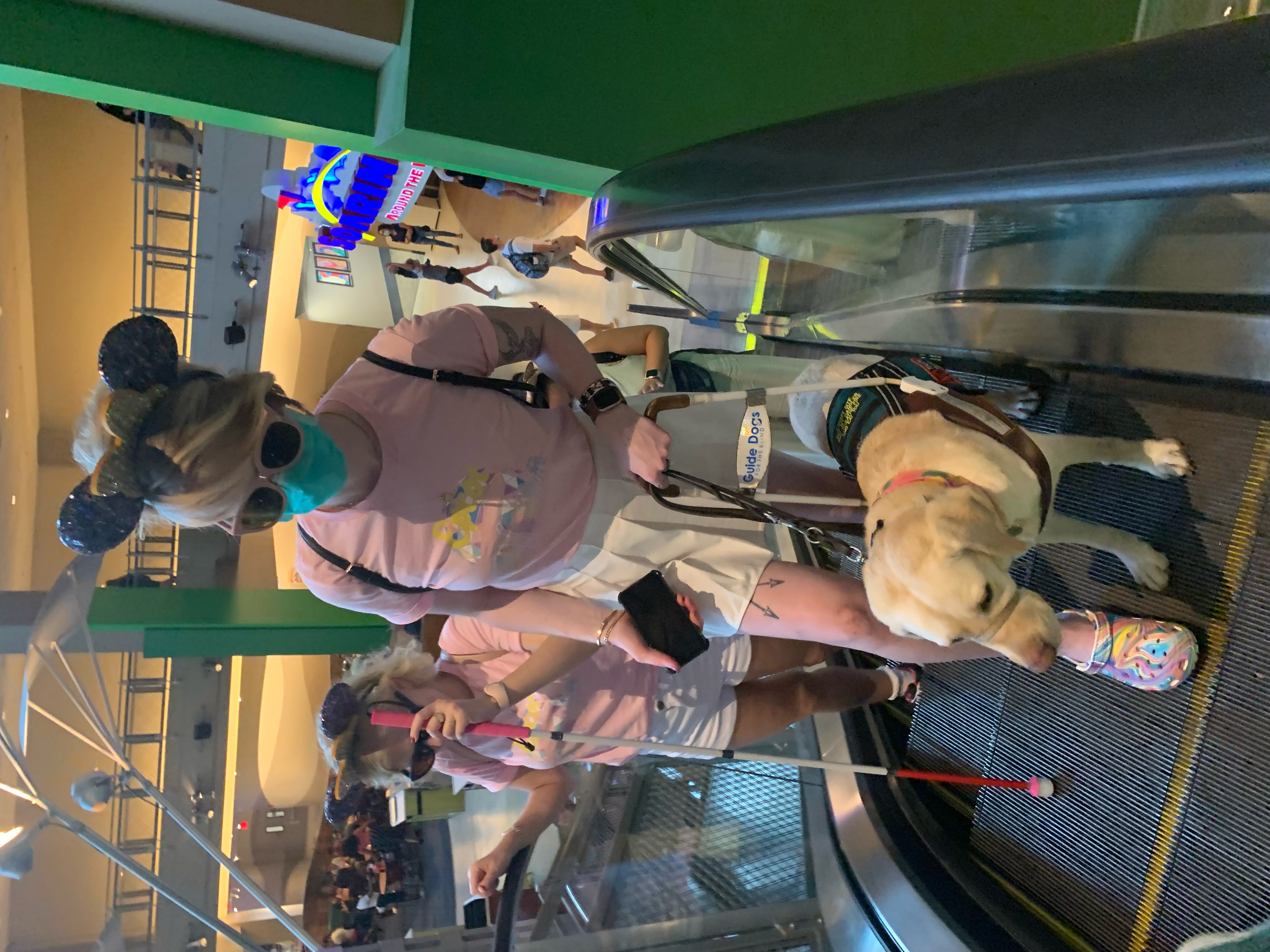 photo of case and cass in disney on an escalator. they wear light pink tshirts and mickey ears. casey uses her hot pink cane and cassandra holds romana's harness handle