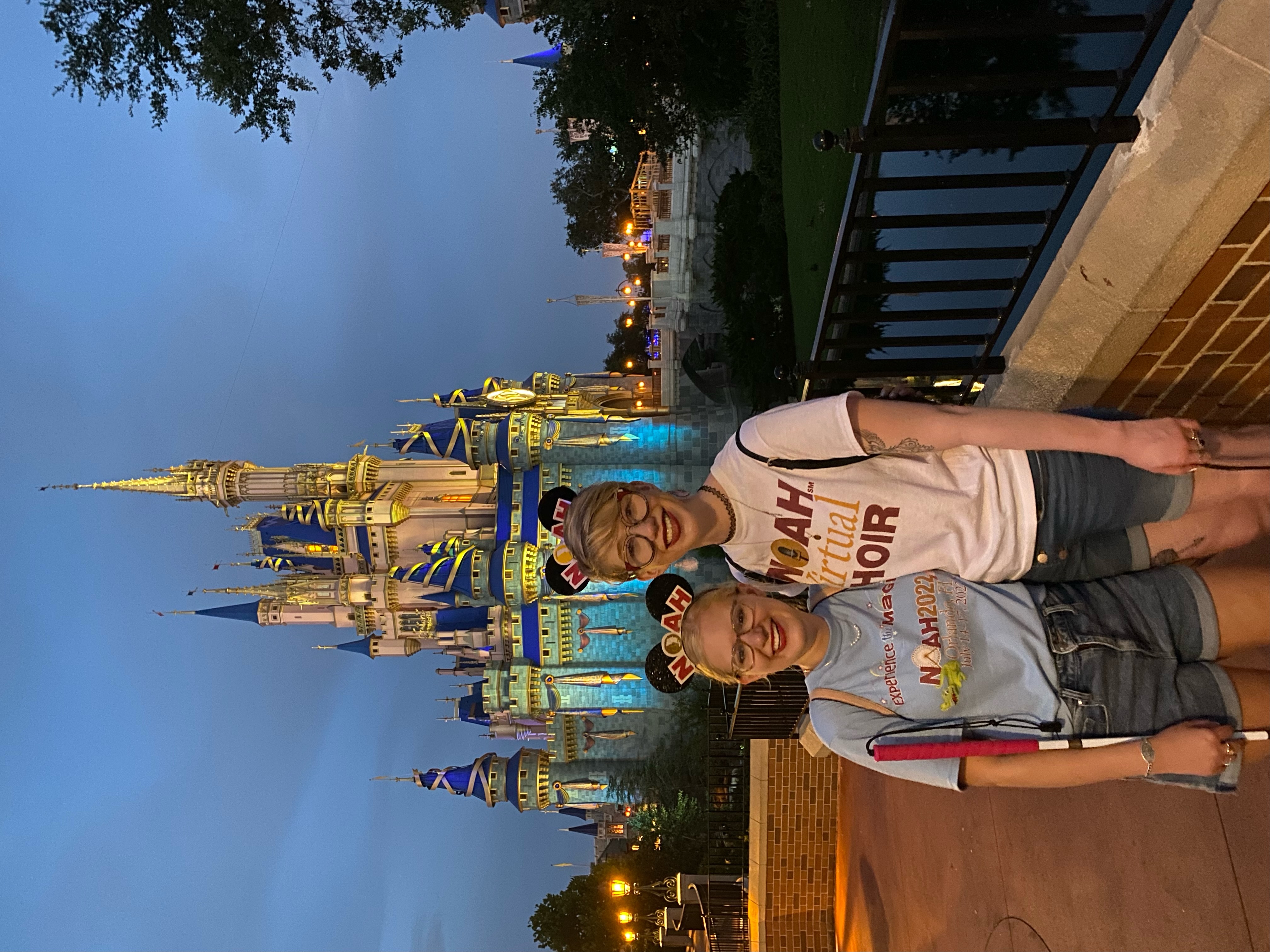 photo of casey and cassandra in front of the cinderella castle at disney world at twilight. they have an arm around each other and wear NOAH t-shirts with NOAH themed Mickey ears. they smile as the castle is cast in light blue spotlights from below
