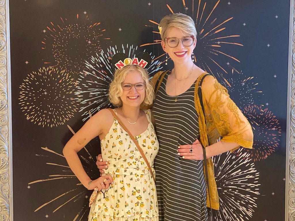 case and cass standing side by side with their arms around each other in front of a backdrop with fireworks. casey wears a white dress with lemons and oranges on it and mickey ears with the NOAH logo. cassandra wears a black and white striped dress, and a yellow cardigan
