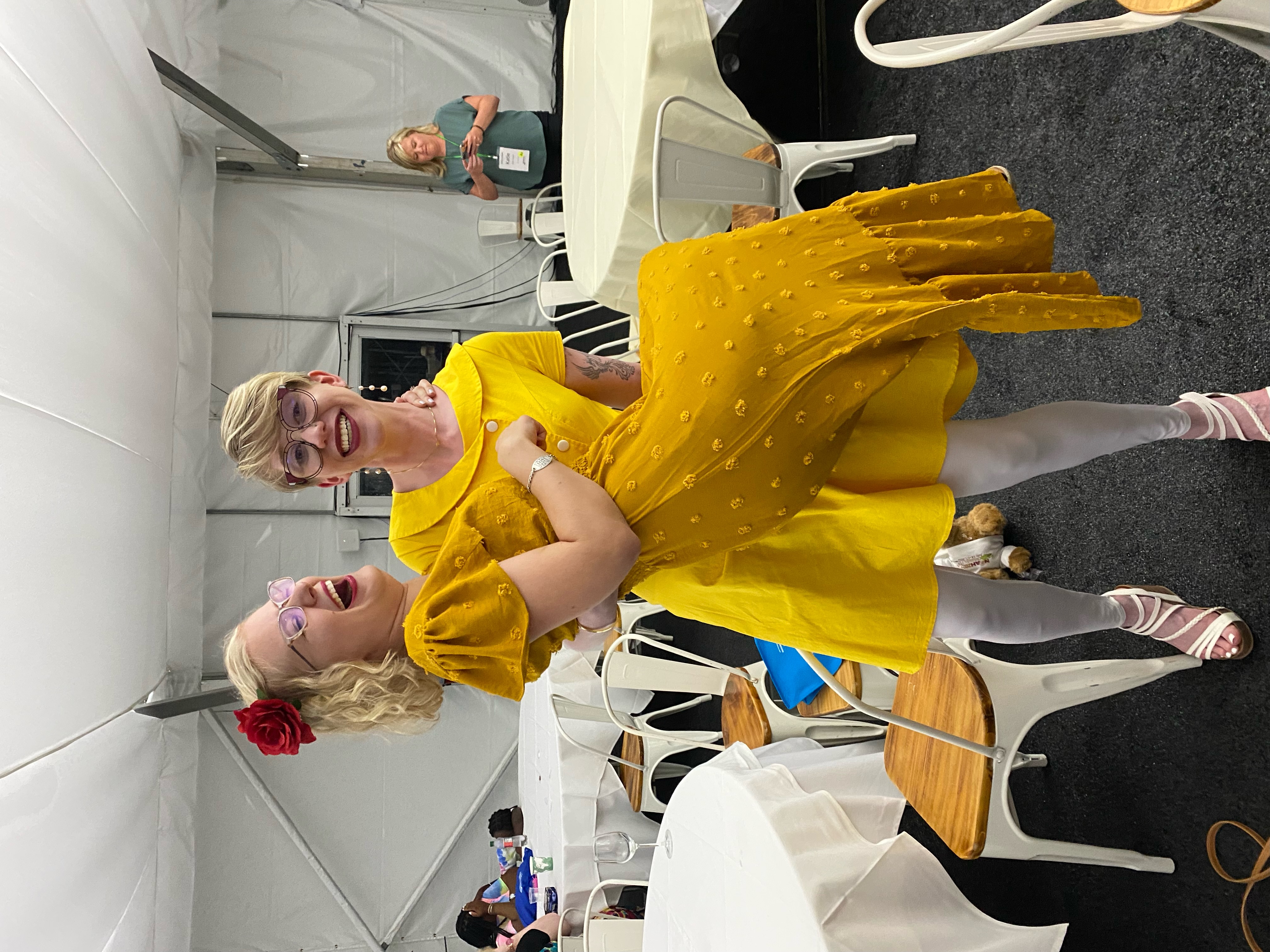 photo of casey and cassandra by some round tables. cassandra wears a yellow dress and takes a wide stance with a goofy grimace as she lifts casey bridal style. casey also wears a yellow dress and laughs at the situation
