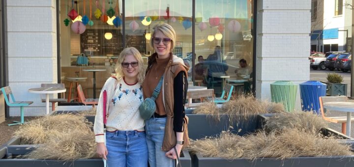 photo of casey and cassandra outside in front of a jenis ice cream shop. case and cass have their arms around each other and smile in the daylight. case holds her pink-handled cane and cass holds yellow lab guide dog romana's leash as romana glances off to the right