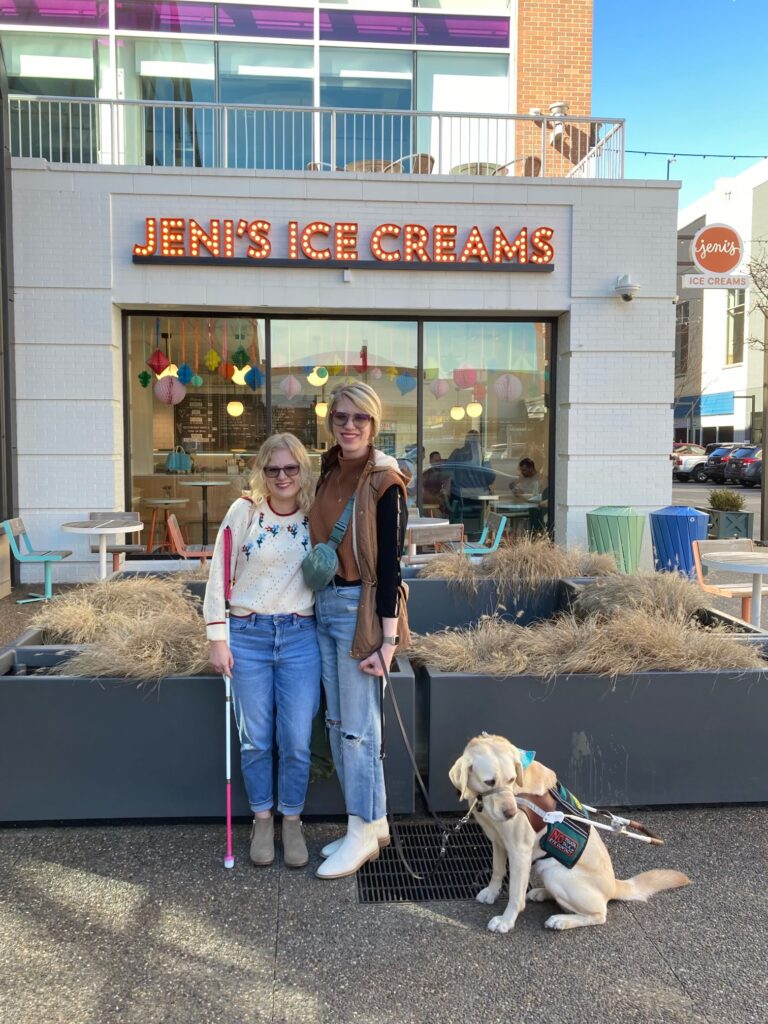 photo of casey and cassandra outside in front of a jenis ice cream shop. case and cass have their arms around each other and smile in the daylight. case holds her pink-handled cane and cass holds yellow lab guide dog romana's leash as romana glances off to the right