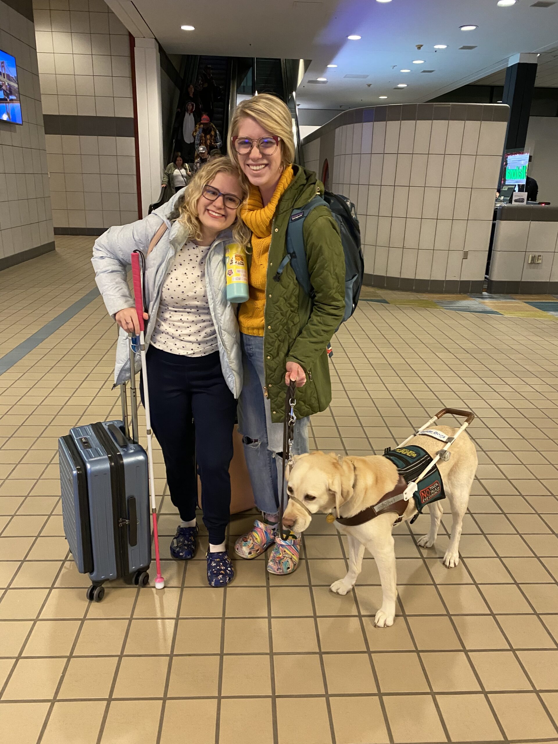 photo of casey and cassandra at an airport. casey, a 5’ blonde with shoulder length hair and glasses l, smiles and holds her pink handled cane and her carry on. cassandra, a 5’9 blonde with a long pixie cut and glasses, stands and grins broadly and stands by her carry on and yellow lab guide dog romana