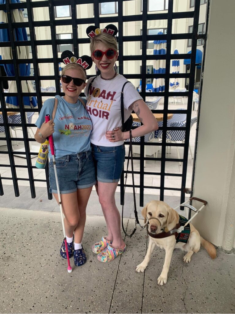 casey and cassandra posing together in the summertime. they both wear national organization for albinism & hypopigmentation shirts and mickey ears and stand outside smiling. they each wear a pair of crocs