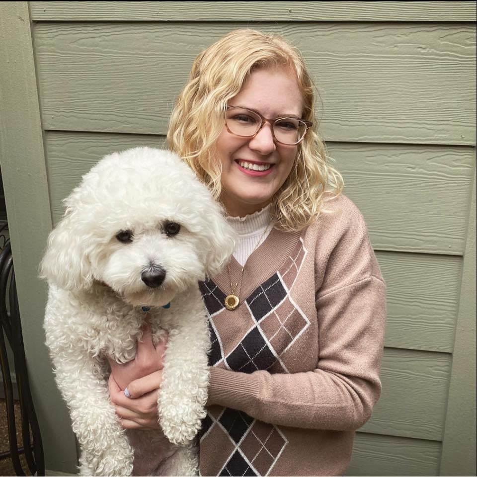 casey, a fair-skinned woman with shoulder-length wavy blonde hair and rectangular glasses, holds her bichon frisé Rupert and smiles