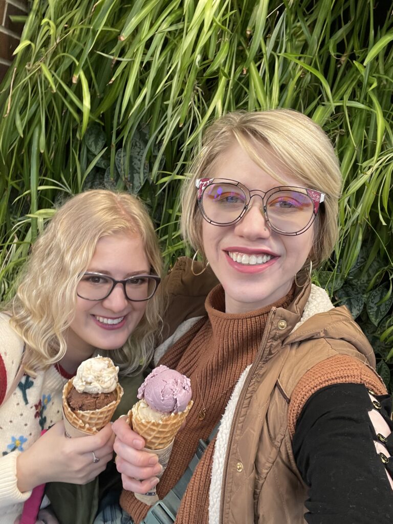 casey and cassandra smile while holding ice cream cones in front of a wall of greenery