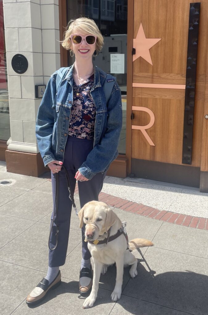 cassandra, a fair skinned woman with chin-length blonde hair, wears pink sunglasses and stands outside with her yellow lab guide dog, romana, sitting at her feet in her harness