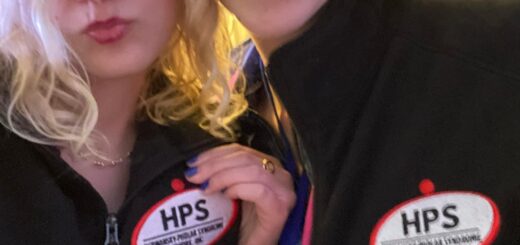 selfie of case, making a duck face, and cass smiling broadly, showing off their matching embroidered vests emblazoned with the HPS network logo and the words "research team"