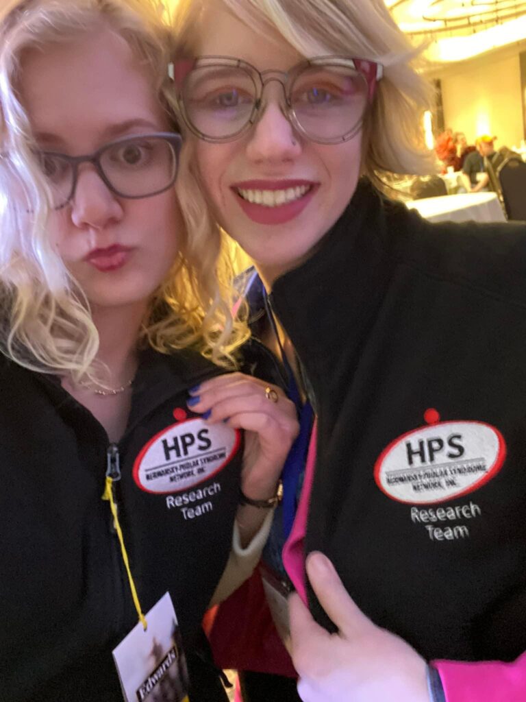 selfie of case, making a duck face, and cass smiling broadly, showing off their matching embroidered vests emblazoned with the HPS network logo and the words "research team"