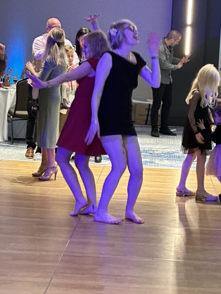 case and cass stand back to back in their party dresses, laughing and singing as they dance on a colorfully lit dance floor