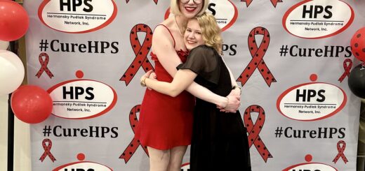 case and cass smile and hug in front of a step-and-repeat background with the HPS Network logo tiled all framed by a collection of red, black, and white balloons, some with polka dots