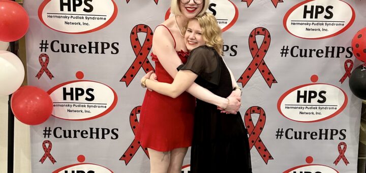 case and cass smile and hug in front of a step-and-repeat background with the HPS Network logo tiled all framed by a collection of red, black, and white balloons, some with polka dots