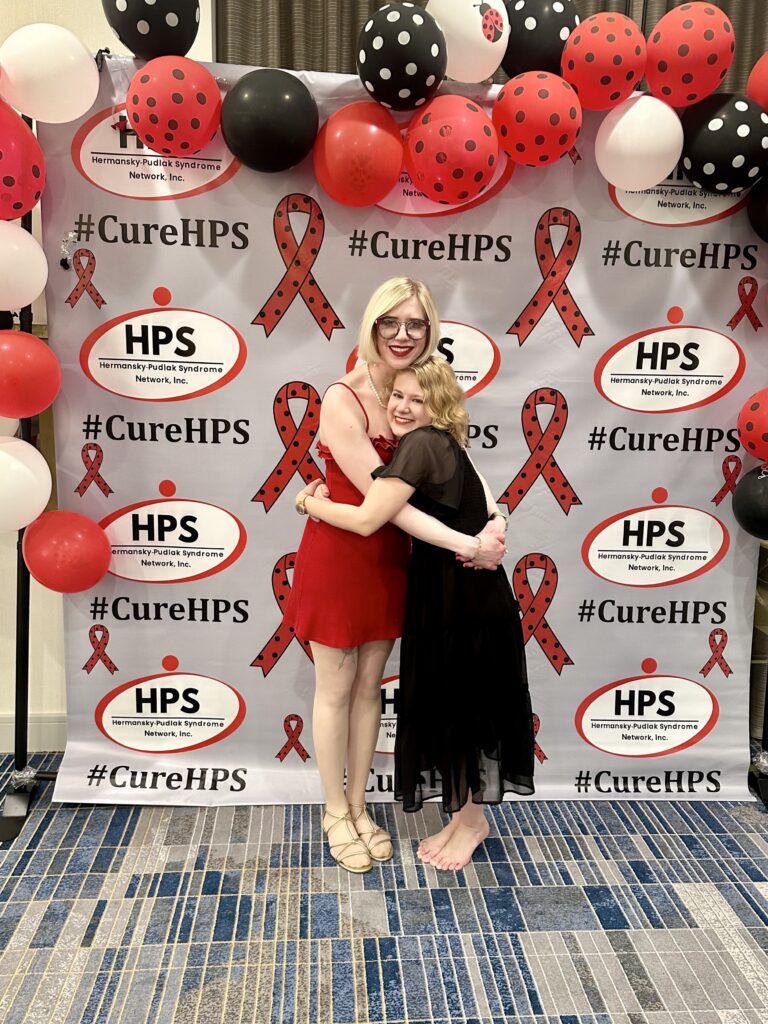 case and cass smile and hug in front of a step-and-repeat background with the HPS Network logo tiled all framed by a collection of red, black, and white balloons, some with polka dots