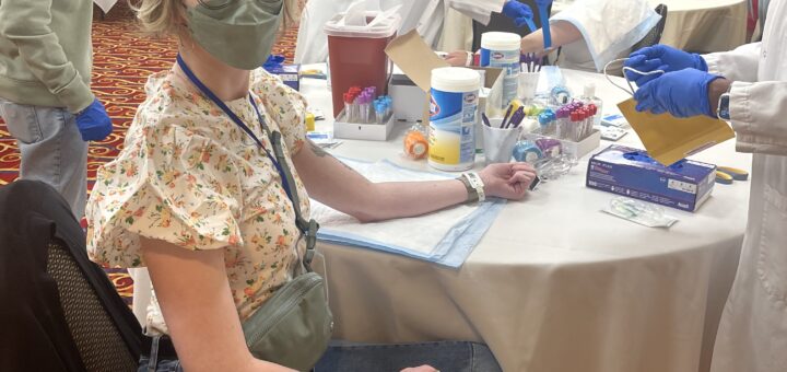 cass and case sit at a table covered in medical supplies in a hotel conference room. they wear masks and extend their arms as healthcare staff draw their blood