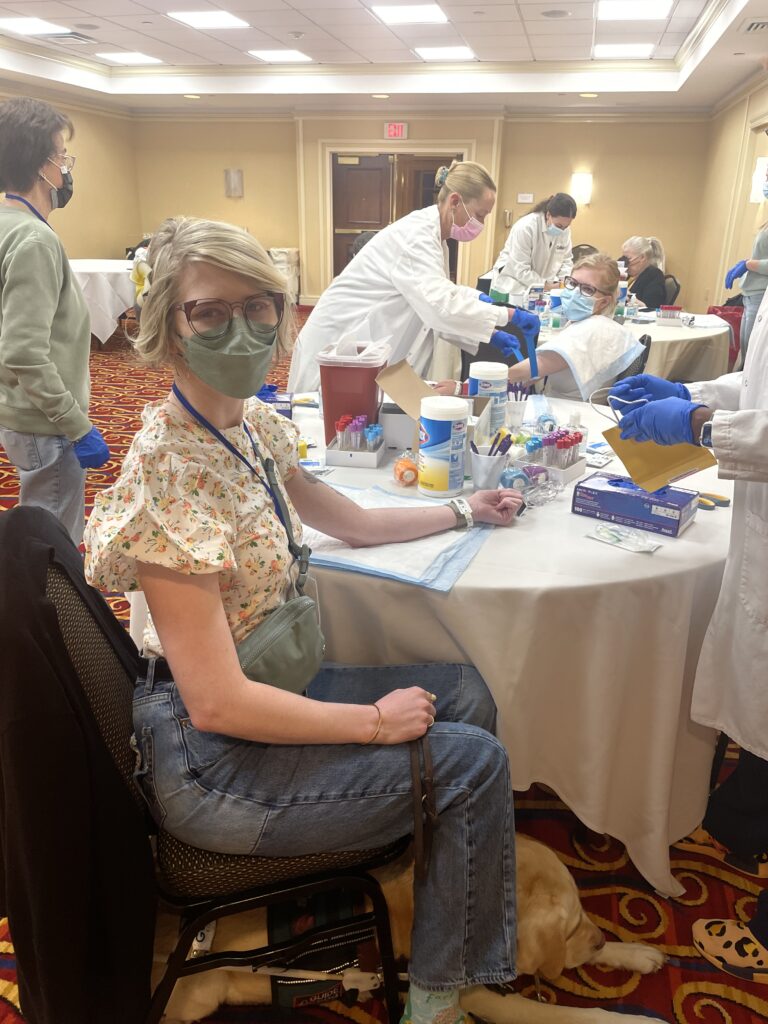 cass and case sit at a table covered in medical supplies in a hotel conference room. they wear masks and extend their arms as healthcare staff draw their blood