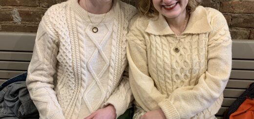 case and cass sit side by side on a bench in a cafe. against an exposed brick wall, the two women smile behind a table with a pastry on a plate. they wear similar cream colored sweaters