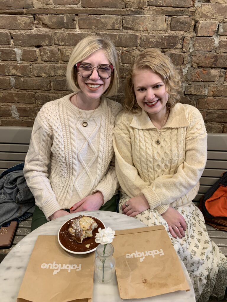 case and cass sit side by side on a bench in a cafe. against an exposed brick wall, the two women smile behind a table with a pastry on a plate. they wear similar cream colored sweaters