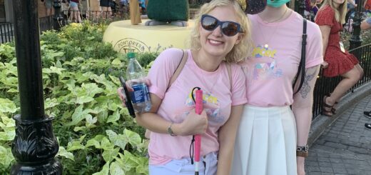 case and cass stand outside a french themed area in disney world. they wear matching pink tshirts and identical bluish purple sequined minnie mouse ears with a big bejeweled yellow bow. both women wear sunglasses and smile joyfully as casey grips her pink handled cane and romana the guide dog stands behind cassandra