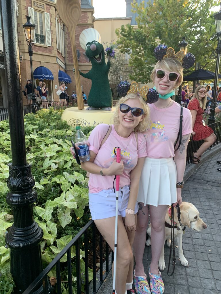 case and cass stand outside a french themed area in disney world. they wear matching pink tshirts and identical bluish purple sequined minnie mouse ears with a big bejeweled yellow bow. both women wear sunglasses and smile joyfully as casey grips her pink handled cane and romana the guide dog stands behind cassandra