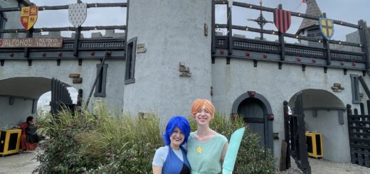 case & cass smile happily in front of a renaissance castle dressed in their steven universe cosplays. case, dressed like lapis lazuli, sports a bright blue wig and blue dress. cass, dressed as pearl, wears a coral wig, mint top, and yellow shorts like the character