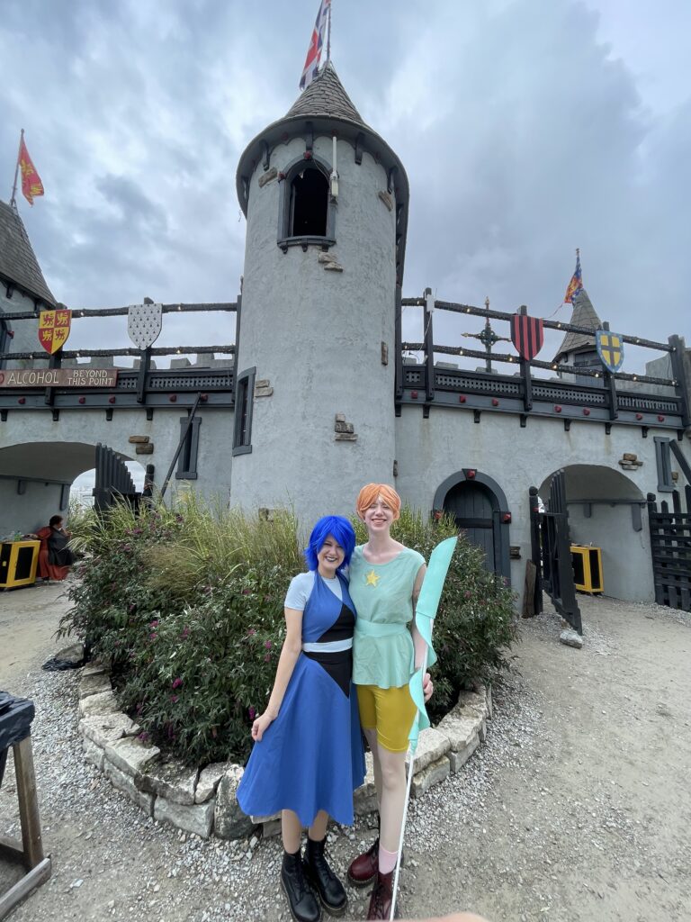 case & cass smile happily in front of a renaissance castle dressed in their steven universe cosplays. case, dressed like lapis lazuli, sports a bright blue wig and blue dress. cass, dressed as pearl, wears a coral wig, mint top, and yellow shorts like the character