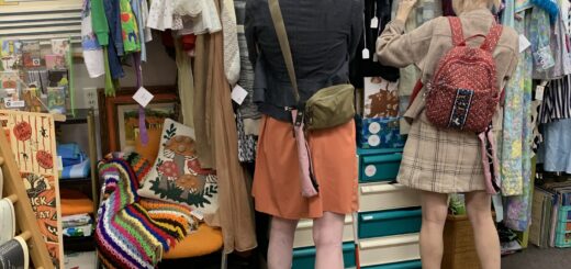 cassandra and casey browse a vintage store. they sift through clothes with their backs to the camera and both of their hairs pulled back and tied with beige bows. their pink cane pouches are visible, attached to each of their bags.