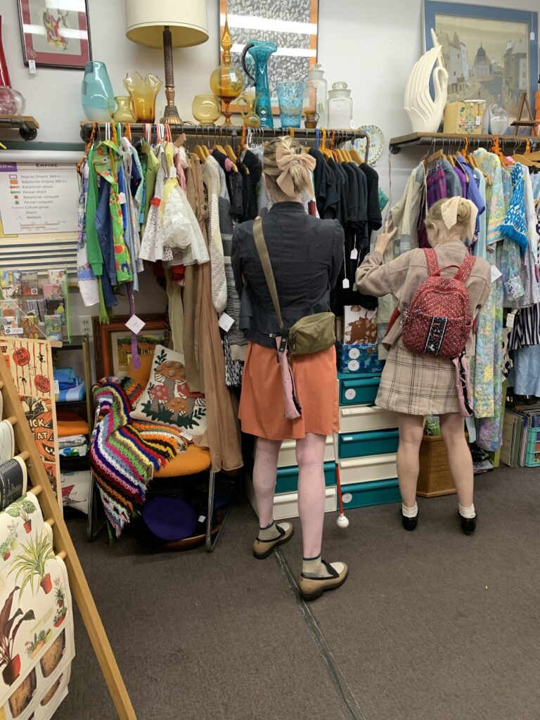 cassandra and casey browse a vintage store. they sift through clothes with their backs to the camera and both of their hairs pulled back and tied with beige bows. their pink cane pouches are visible, attached to each of their bags.